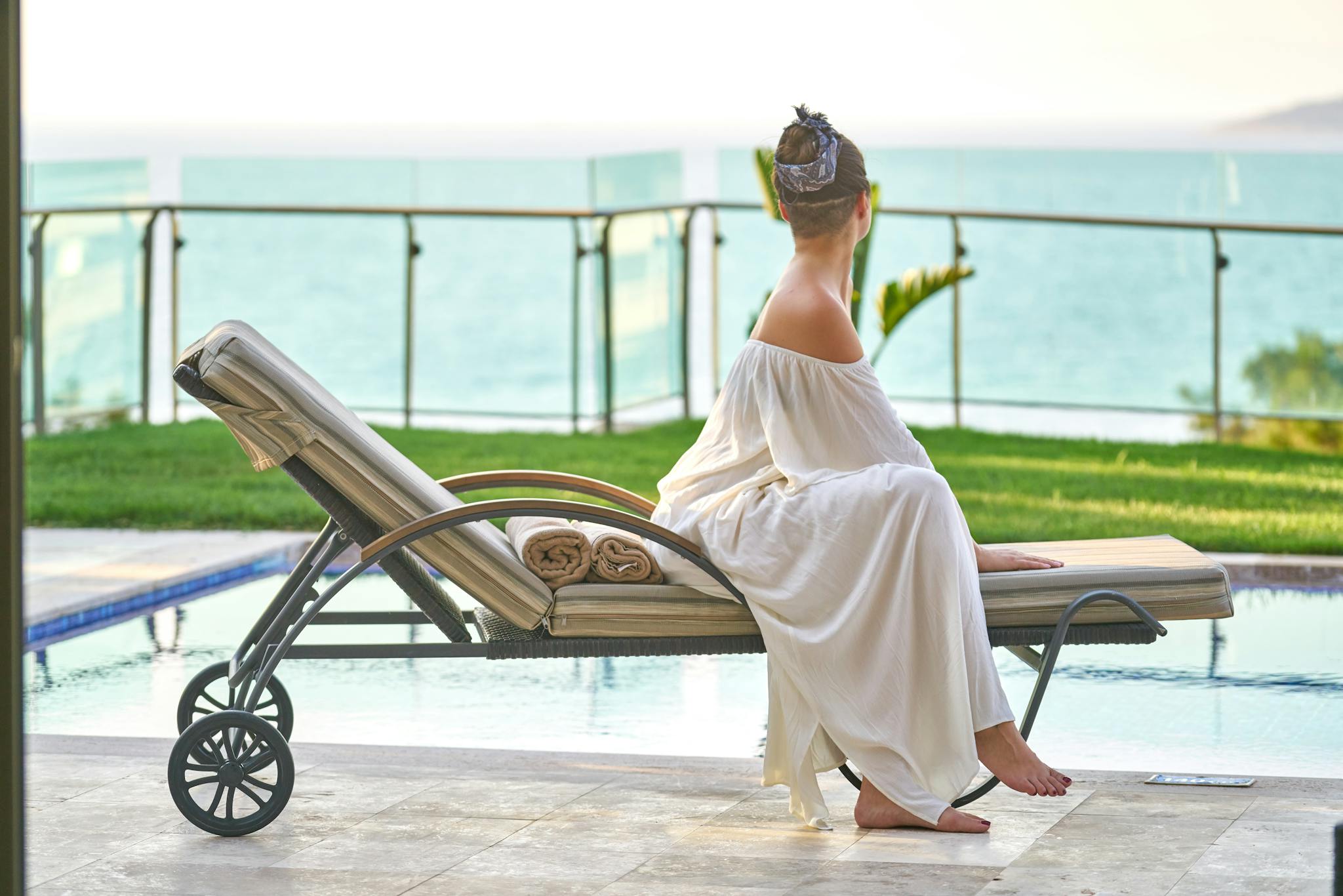 A woman in a white dress relaxes by the poolside with an ocean view.